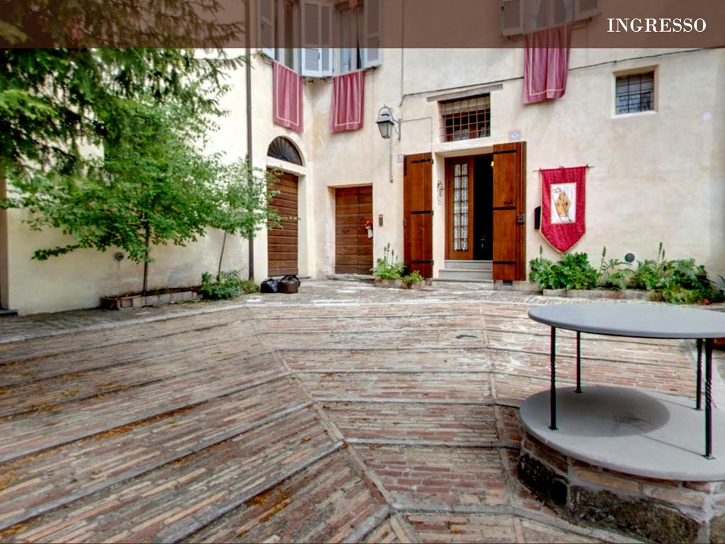 The entrance area features a rustic courtyard paved with brick, bordered by greenery and plants. Wooden doors, flanked by decorative banners, lead into the building. A simple circular table is positioned on one side, providing a space for relaxation.