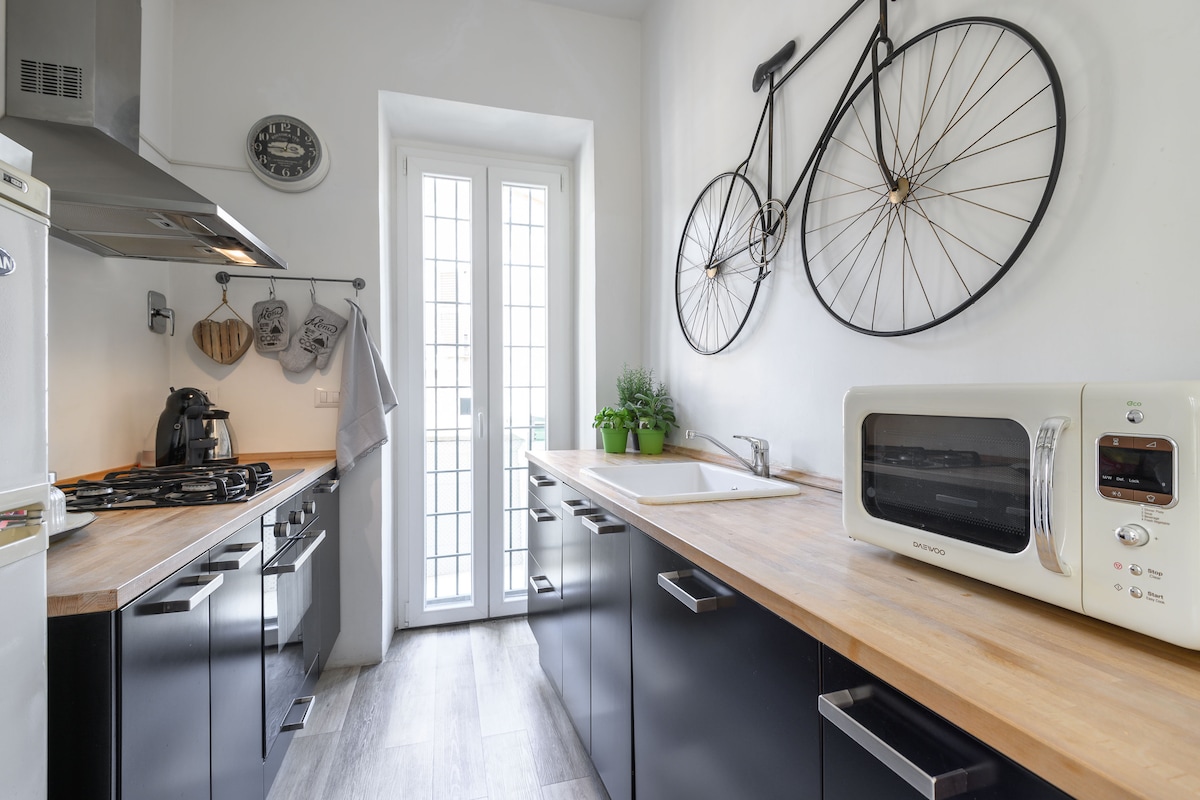 A modern kitchen is presented with dark cabinetry and a light wood countertop. Decorative bicycle wheels hang on the wall, adding character. A window provides natural light, and appliances include a gas stove, microwave, and a sink. Potted plants sit on the counter, enhancing the space.