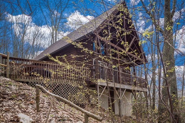 A rustic cabin is framed by trees and greenery, showcasing a sloped wooden deck with railings. The structure features a steeply pitched roof and large front windows that invite natural light. Soft clouds float in a bright blue sky.