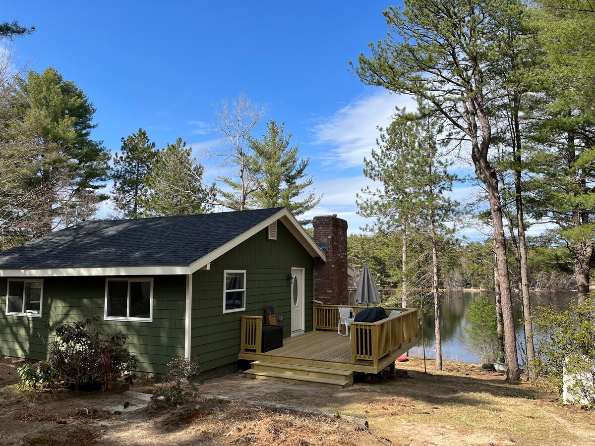 A quiet green cottage is pictured beside a serene lake, surrounded by trees. A wooden deck is shown in front, featuring seating and a grill. The blue sky is visible above, reflecting the peaceful atmosphere of the natural setting.