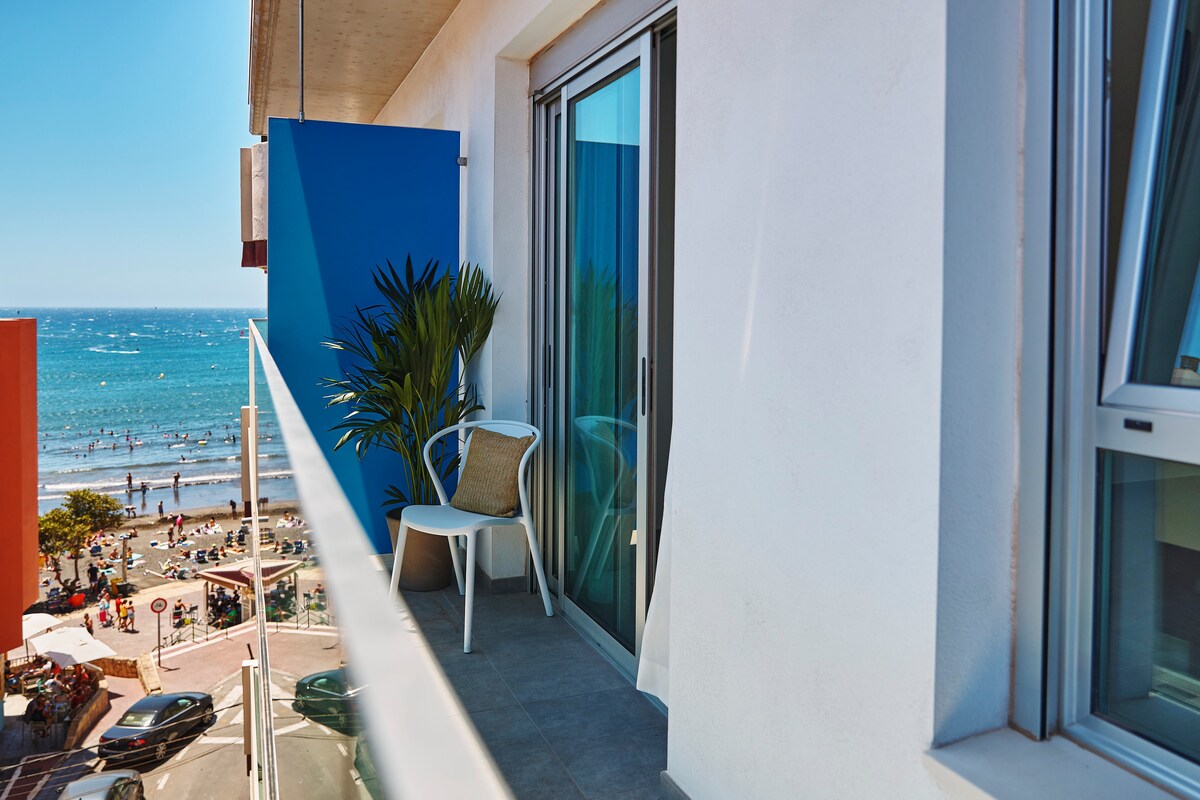 A balcony is shown with a clear view of the sea and sandy beach below. A single chair is placed beside a potted plant against a vibrant blue wall. Large glass doors lead inside, allowing natural light to enhance the space.