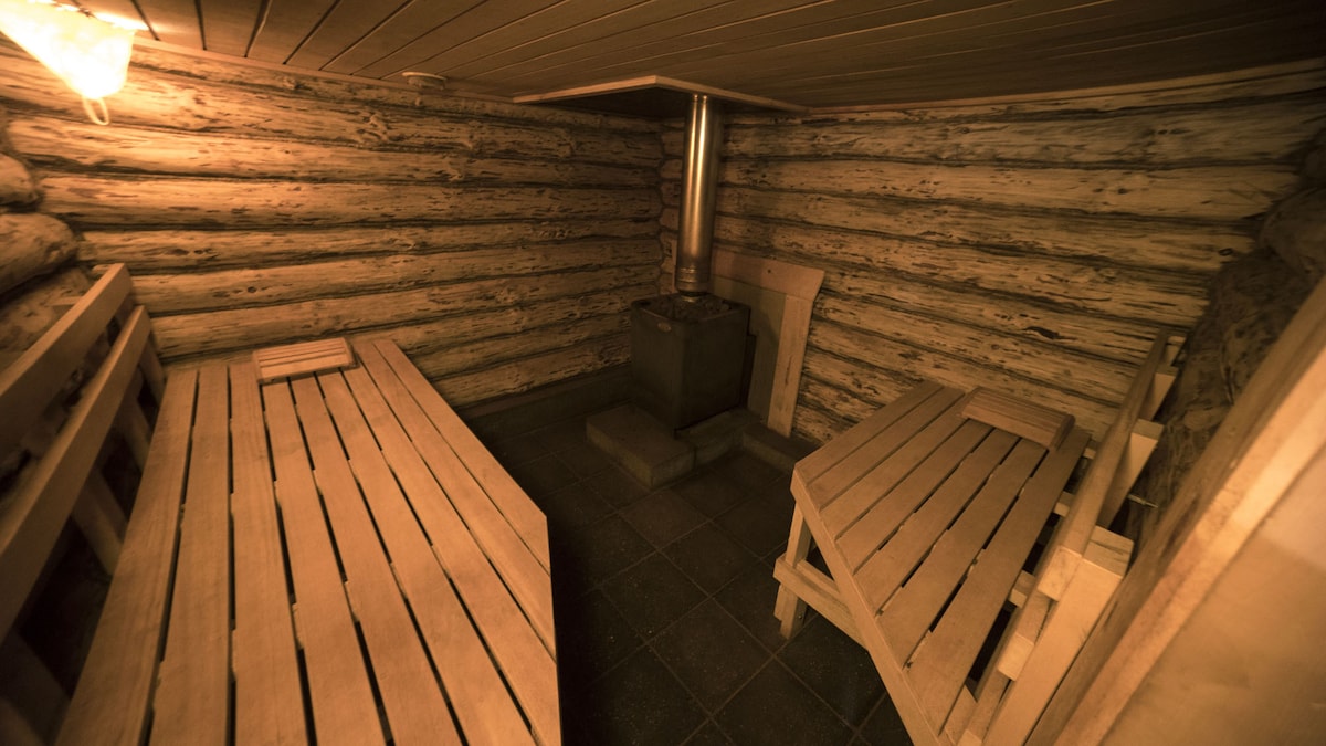 A rustic steam bath is seen, featuring wooden benches arranged around a central stone heater. The walls are made of stacked logs, and the floor is tiled. Soft lighting creates a warm ambiance, inviting guests to relax in the serene environment.