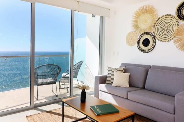 A modern living space features large glass doors that open to a private terrace overlooking the ocean. A gray sofa is positioned beside a coffee table, with natural elements like a decorative vase and wall art adding texture to the room.