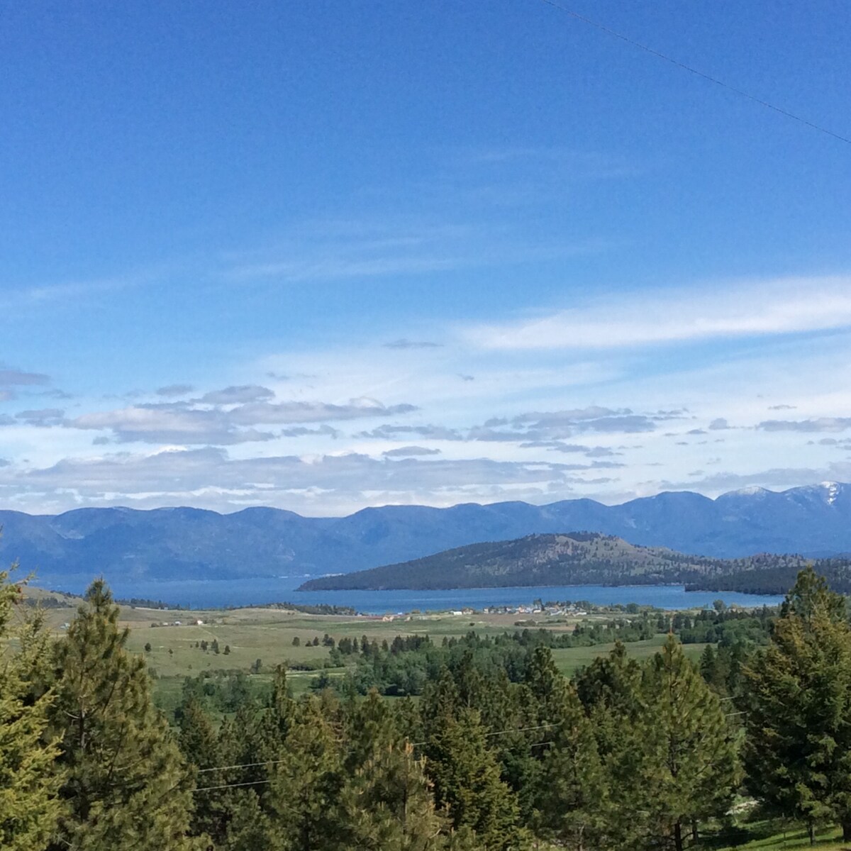 A panoramic view showcases rolling hills and a shimmering lake, framed by majestic mountains in the distance. Lush greenery and clusters of trees are visible in the foreground, enhancing the serene landscape under a clear blue sky.