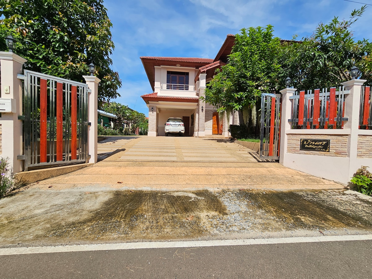 The entrance features a large driveway leading to a two-story house, framed by decorative gates. Lush greenery and trees are visible along the driveway, providing shade and a welcoming atmosphere. A parked car is positioned on the gravel surface, adding to the property's spaciousness.
