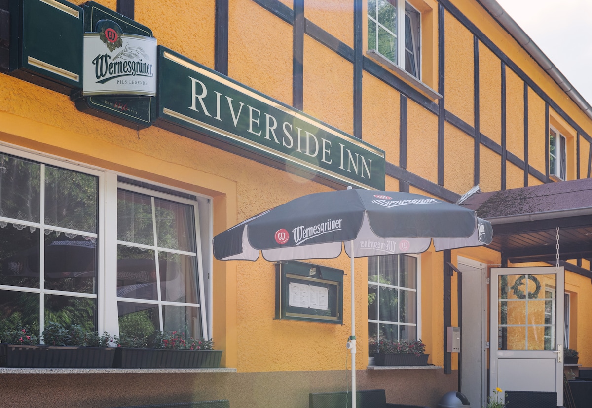 The exterior of the Riverside Inn is showcased, featuring a vibrant yellow facade and a sign that identifies the establishment. A black umbrella is positioned over a small table outside, and there are window boxes with plants below the windows, enhancing the inviting appearance.