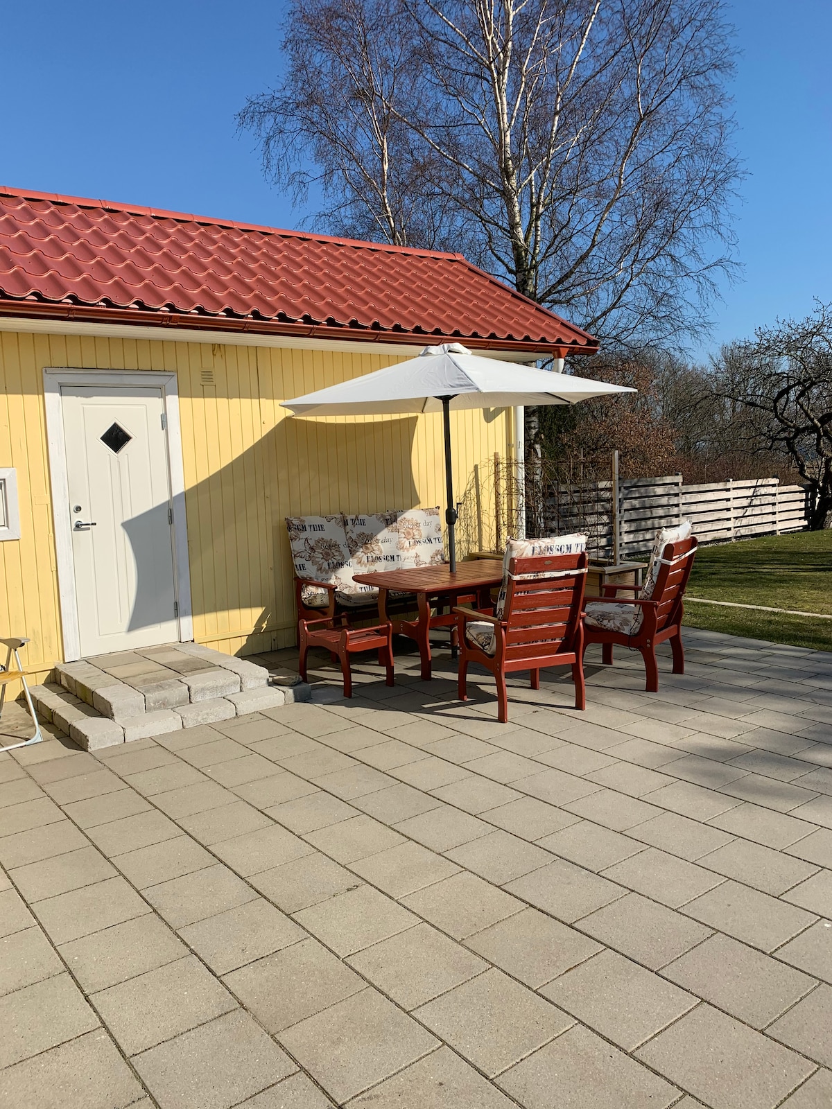 An outdoor patio area features a wooden table accompanied by four high-backed chairs, shaded by a large umbrella. The space is paved with light-colored stones and is adjacent to a well-maintained lawn, with a vibrant yellow exterior of the cottage in the background.