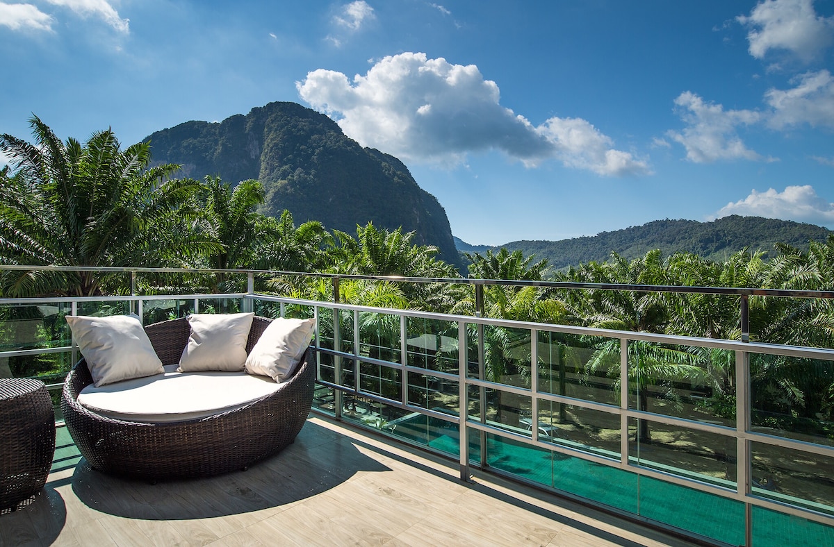 A terrace seating area features a round wicker chair adorned with soft cushions, providing a comfortable spot to relax. Lush greenery and a mountain landscape create a serene backdrop, while a clear blue sky accentuates the vibrant surroundings.