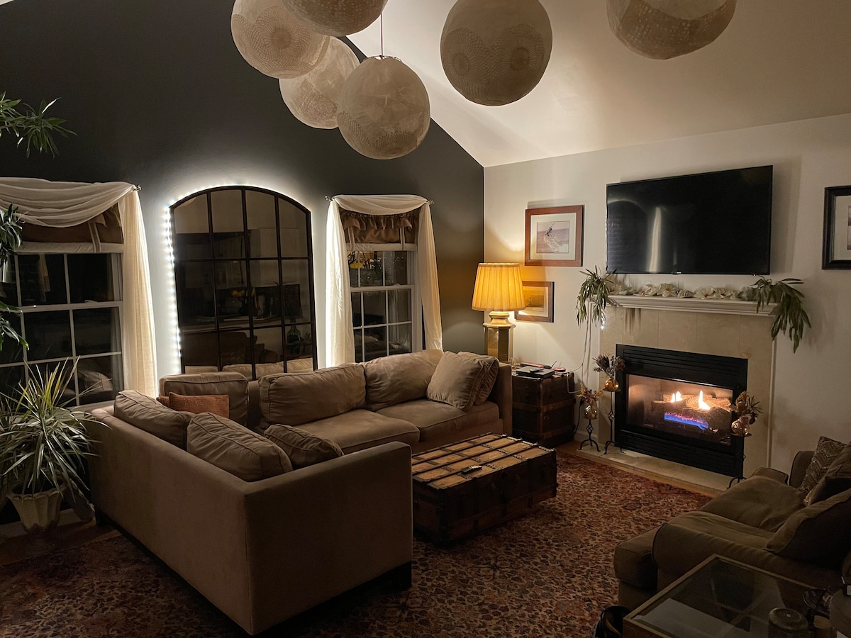 A cozy living room features a large sectional sofa arranged around a wooden coffee table. Illuminated by warm lighting, the space is adorned with plants and a decorative area rug. A fireplace with a modern insert adds to the inviting ambiance, while a flatscreen TV is mounted on the wall.