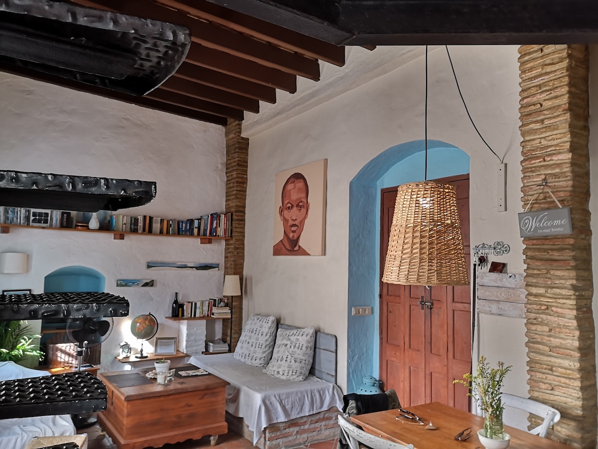A cozy living area features a mix of seating options with two built-in sofas and a wooden coffee table. Shelves lined with books are visible against one wall, while a woven light fixture hangs above a wooden dining table. An arched doorway leads to the entrance.
