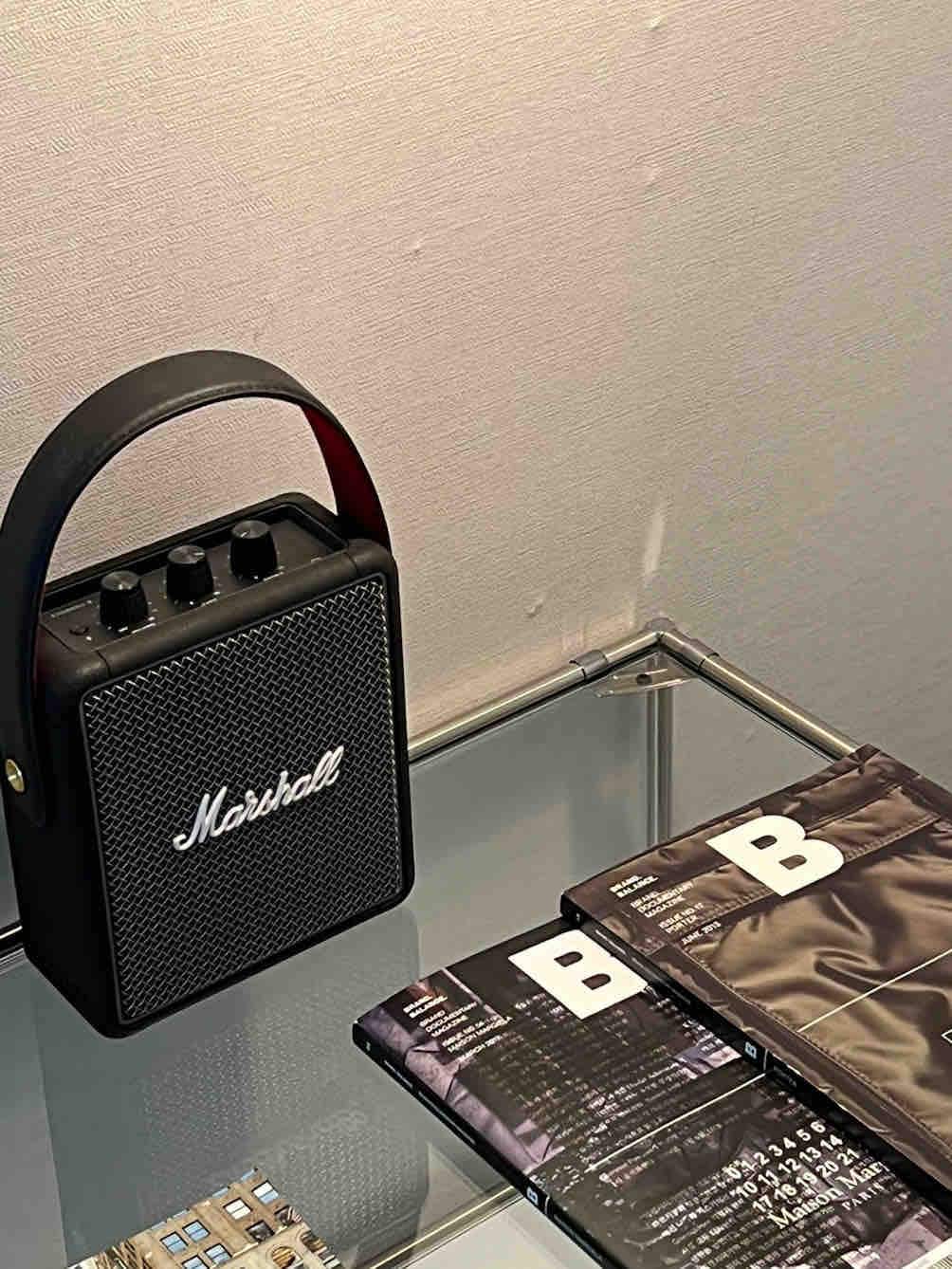 A compact Marshall speaker is positioned on a glass surface alongside several glossy magazines. The minimalist setup showcases a seamless blend of modern design and entertainment elements.