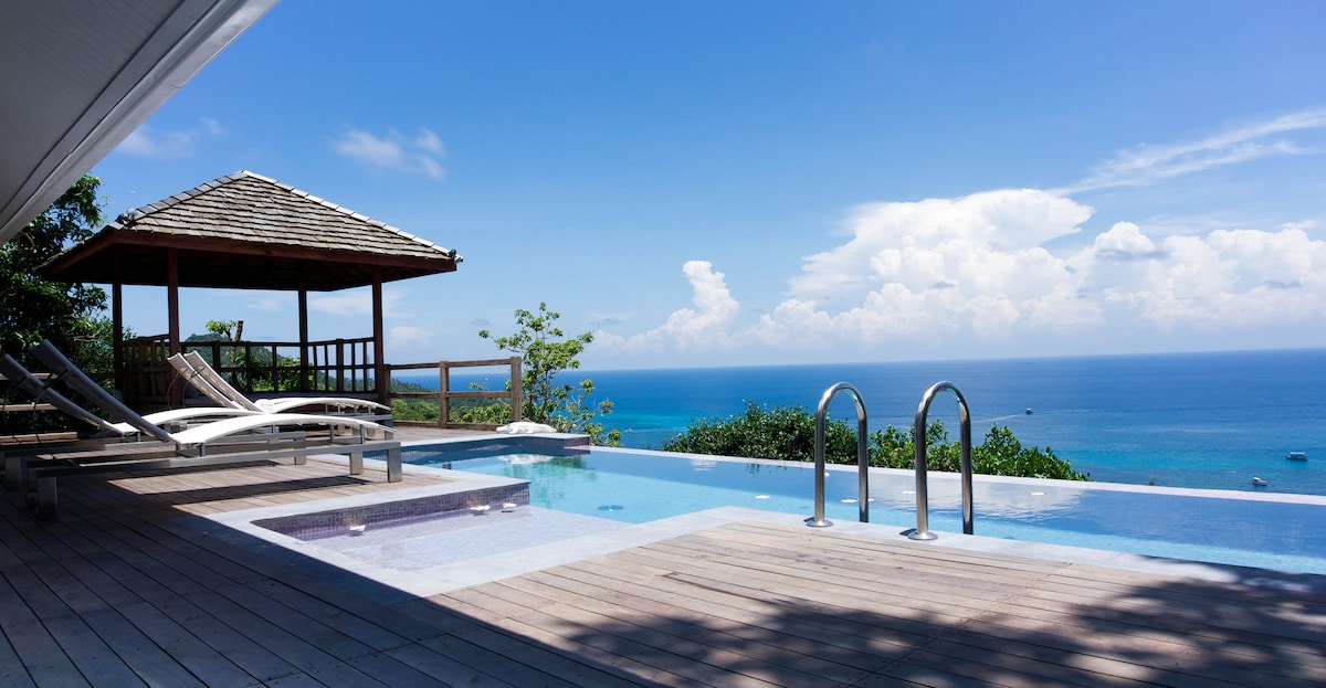 An infinity pool with railing extends towards panoramic ocean views. A wooden gazebo is situated beside the pool, providing shaded seating. Sun loungers are arranged nearby, overlooking the sea, with lush greenery bordering the area. A clear blue sky adds to the tranquil environment.