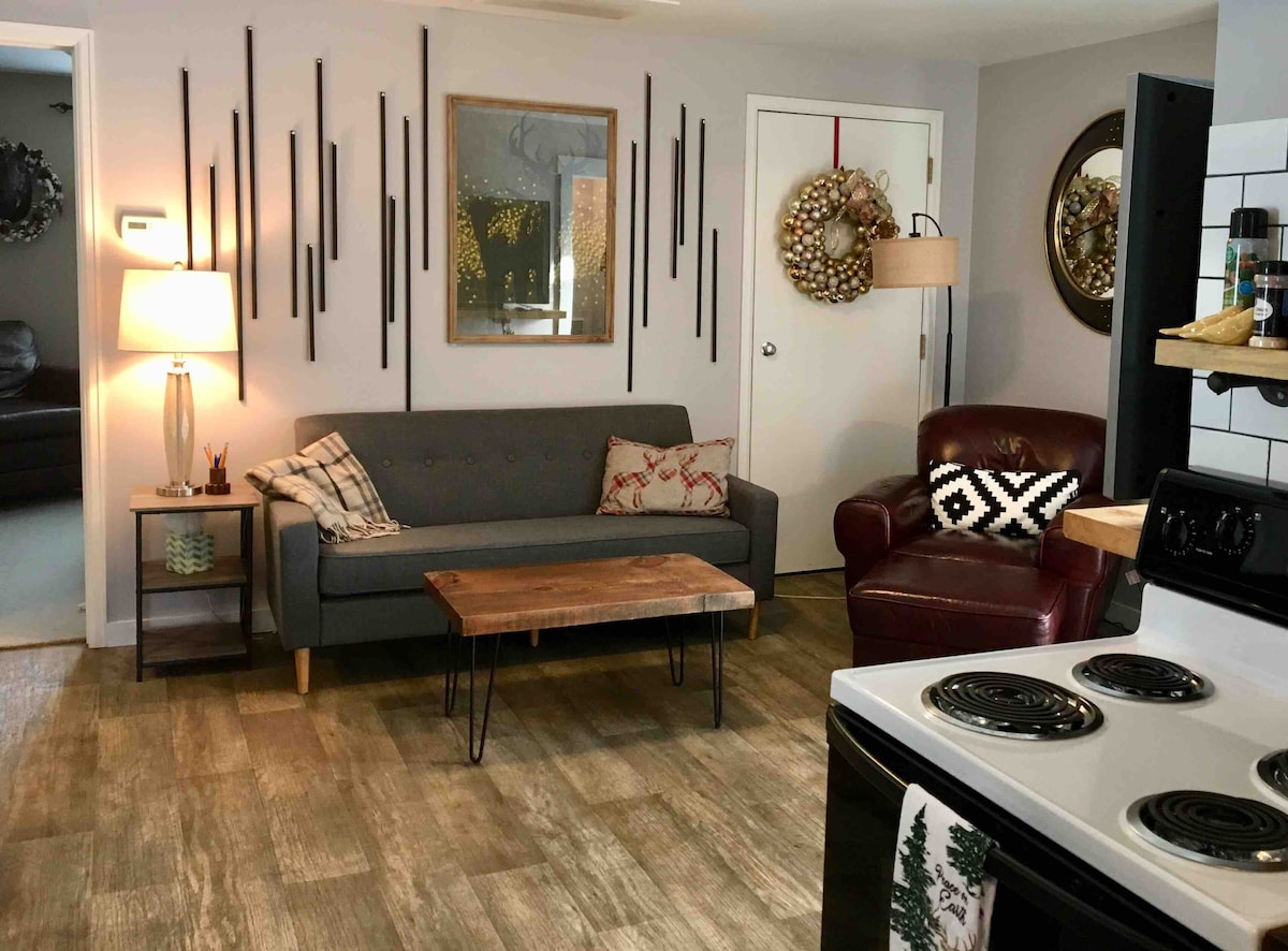 A cozy living area features a gray sofa with a textured throw and a rustic wooden coffee table. A red armchair complements the space, while wall art and a wreath add character. A lamp provides warm lighting, and a kitchen stove is visible in the foreground.
