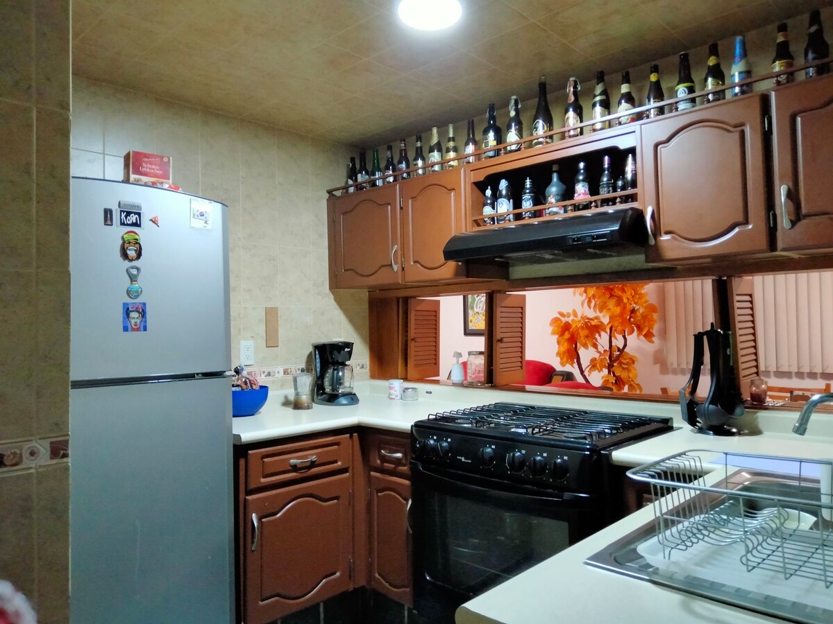 A functional kitchen is equipped with essential appliances, including a refrigerator, stove, and coffee maker. Decorative bottles line the shelves above, contributing a personal touch. Ample counter space is available for meal preparation, and a sink is positioned conveniently near the cooking area.