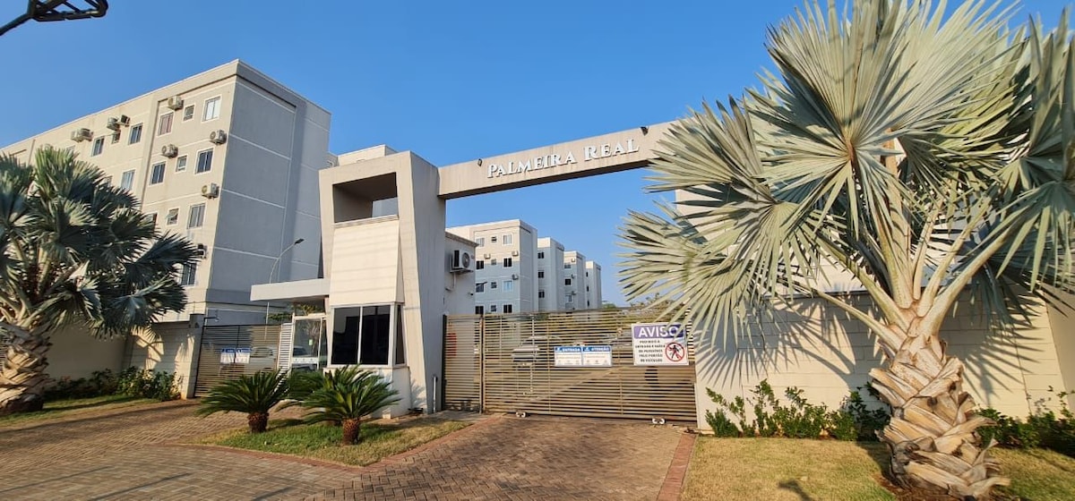 A gated entrance is framed by palm trees, leading to a multi-story building complex. The clear blue sky above enhances the vibrant atmosphere. The entrance features an arch sign that reads 'Palmeira Real,' with a manicured lawn and paved pathway visible in the foreground.