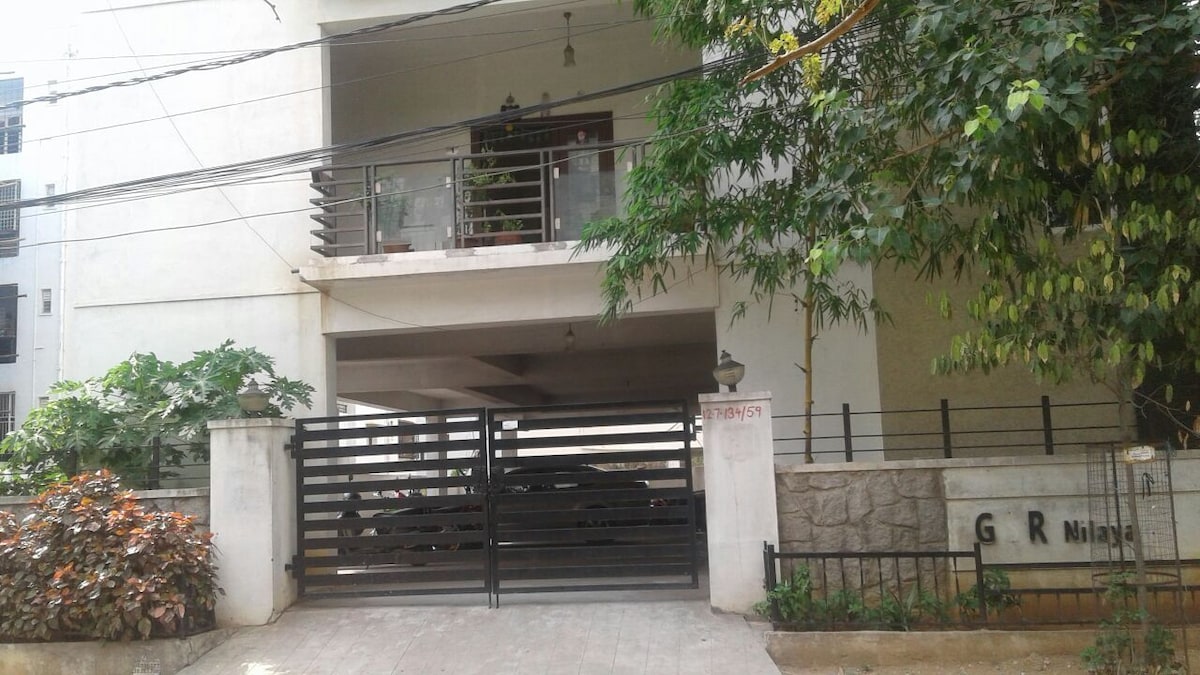 The building exterior features a modern design with a gated entrance and an upper balcony. A parking area can be seen beneath, sheltered from the elements. Lush greenery surrounds the property, adding a touch of nature to the urban setting.