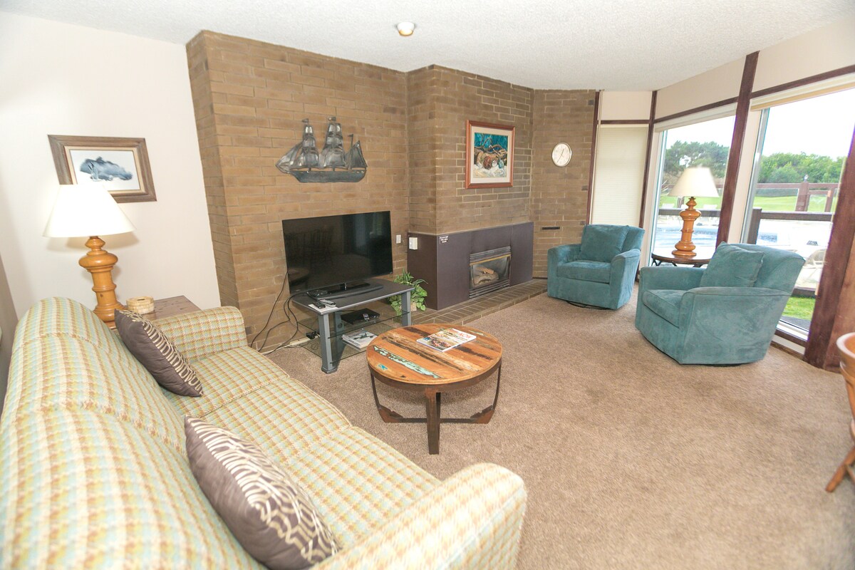 A comfortable living area is presented with a light brown carpet and brick accent wall. A green couch and two blue armchairs are arranged around a wooden coffee table. A flat-screen TV is positioned beside a gas fireplace, with natural light filtering in through large windows.