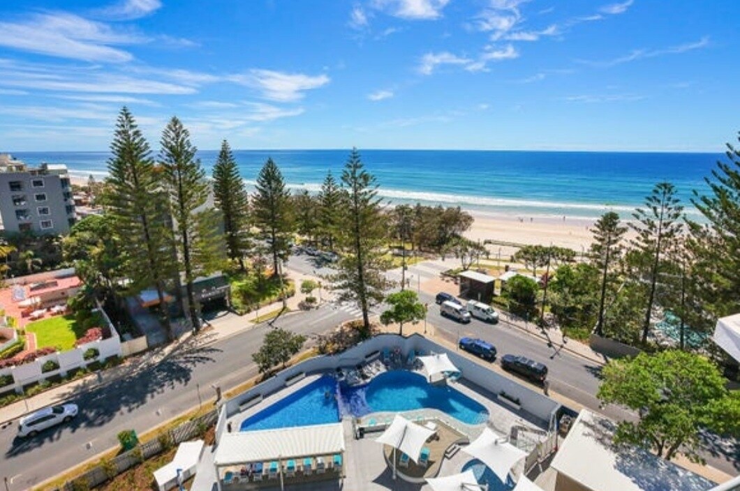 A panoramic view captures the sparkling ocean with soft waves, framed by tall palm trees along the shoreline. The vibrant pool area is visible below, surrounded by sun loungers and white canopies, blending seamlessly with the coastal landscape.