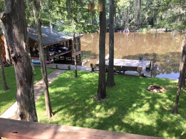 A serene view of a well-maintained lawn leads to a boat house and dock along the calm waters of Caddo Lake. Towering trees provide shade, with a fire pit positioned on the grassy area, enhancing the peaceful outdoor setting.