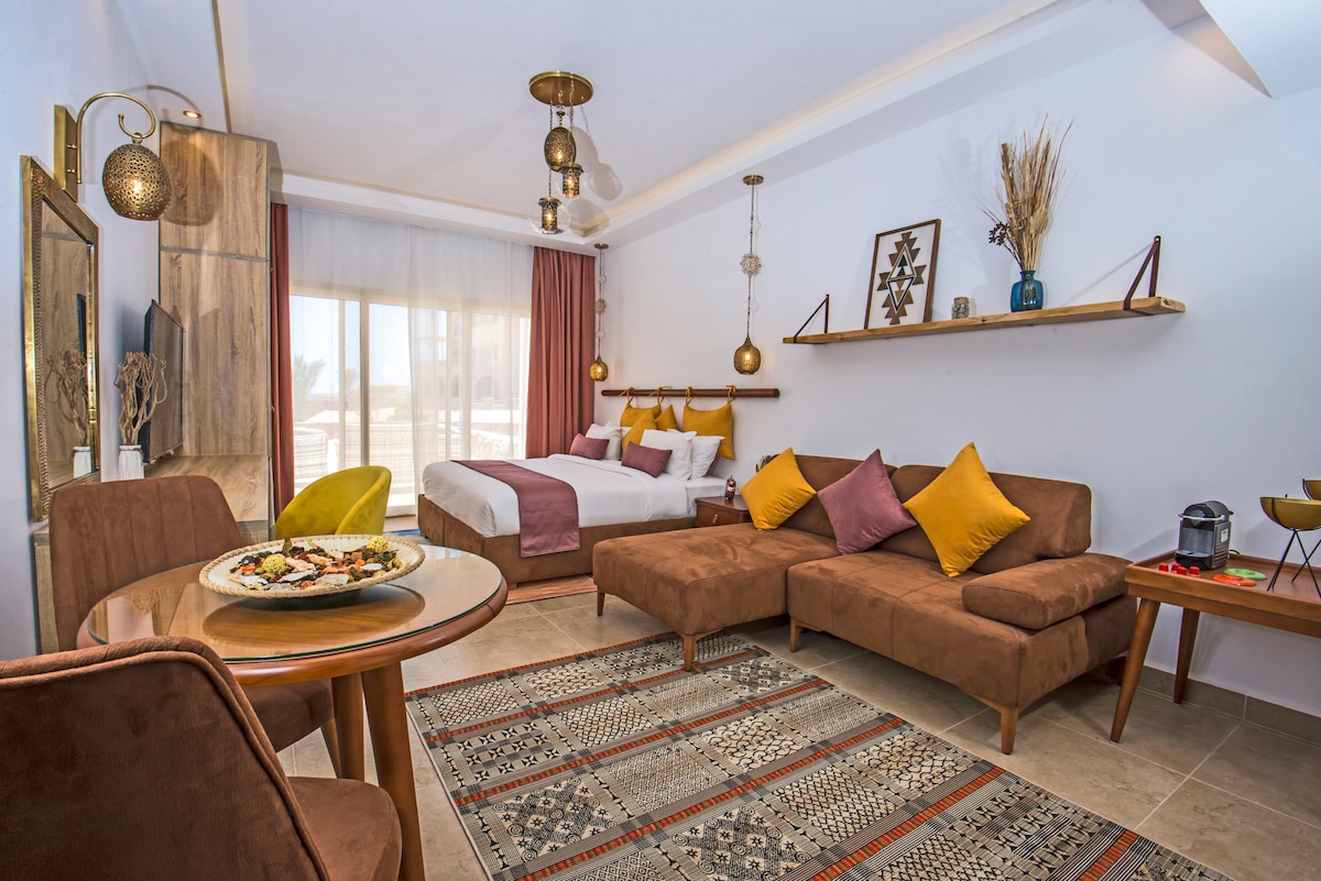 A studio apartment featuring a large bed with decorative pillows and a cozy seating area. A round dining table sits in front of a plush sofa, while light filters through large window drapes. Decorative accents include wall art and a shelf with plants.