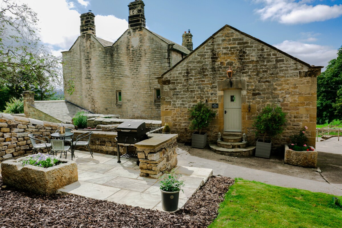 An exterior view of The Old Cooling House features a charming stone facade with a light-colored door. A spacious patio area is seen in the foreground, furnished with a dining set, BBQ grill, and potted plants. Lush greenery and pathways surround the building.
