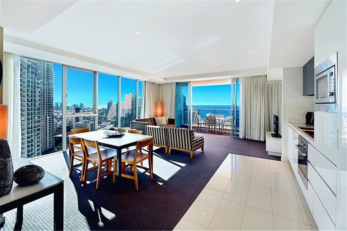 An open living area features floor-to-ceiling windows displaying stunning ocean and city views. A dining table is set for four, while a comfortable seating area with a striped couch is located nearby. Natural light fills the space, complementing the modern kitchen and elegant décor.