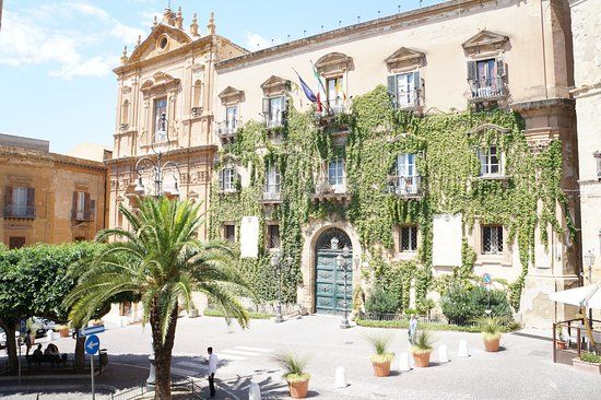 An elegant historical building is highlighted, adorned with lush green ivy. Large windows with balconies are visible on the façade, and a grand entrance is framed by decorative details. Surrounding palm trees and potted plants enhance the inviting atmosphere of the square.