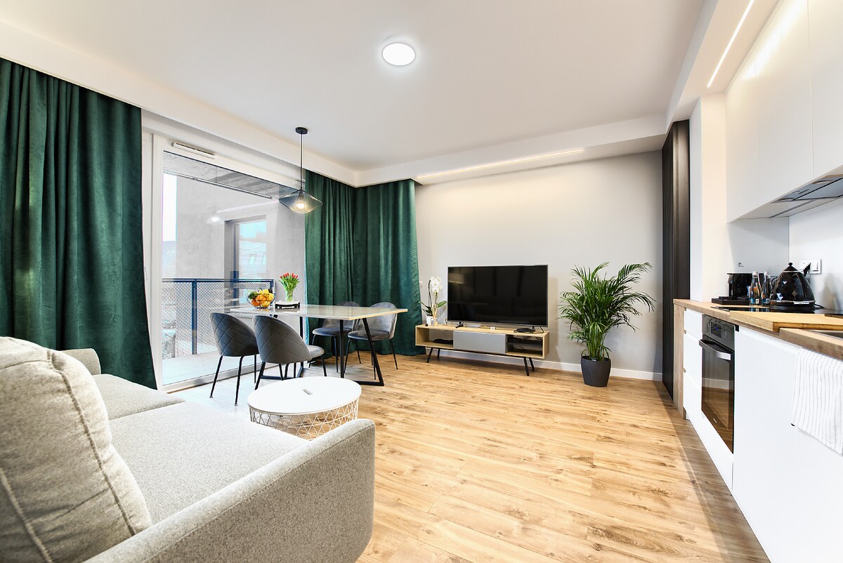 A light-filled living area features a modern sofa and a dining table with four chairs. Large curtains frame the floor-to-ceiling windows that provide access to a balcony, while a television sits on a low media console. A potted plant adds a touch of greenery.
