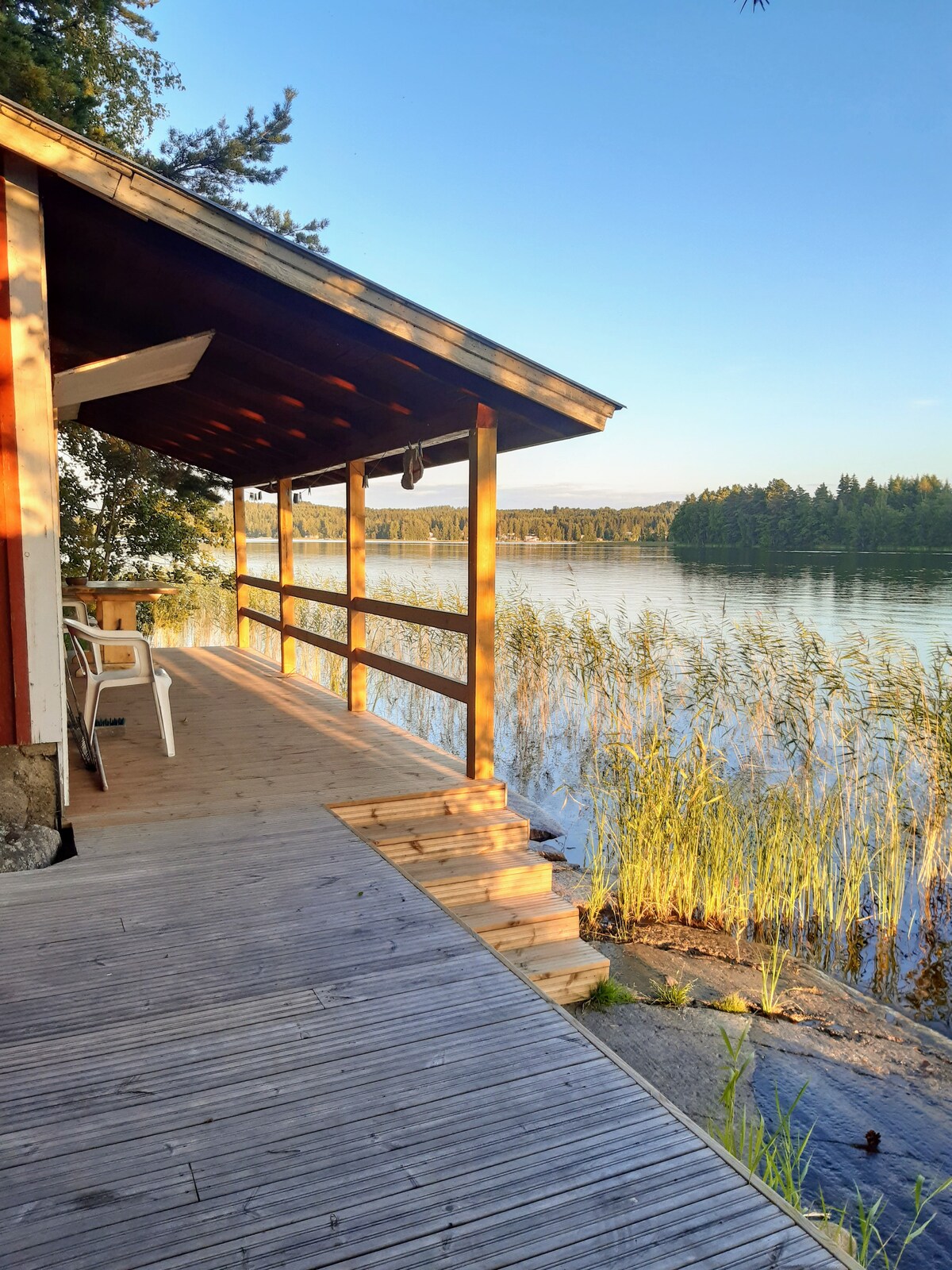 A wooden terrace extends from the summer cottage, overlooking a serene lake. Gentle reeds sway in the water, while trees form a natural backdrop. The space includes a small table and chairs, inviting relaxation by the lakeside.