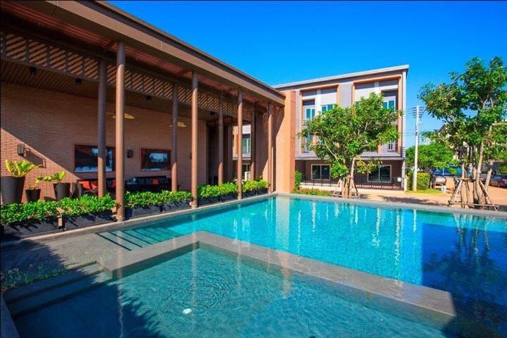 A serene pool area surrounded by lush greenery and modern architecture is visible. The pool features a clear turquoise surface, reflecting the bright sky. Adjacent seating areas are shaded by wooden overhangs, providing space for relaxation and gatherings.