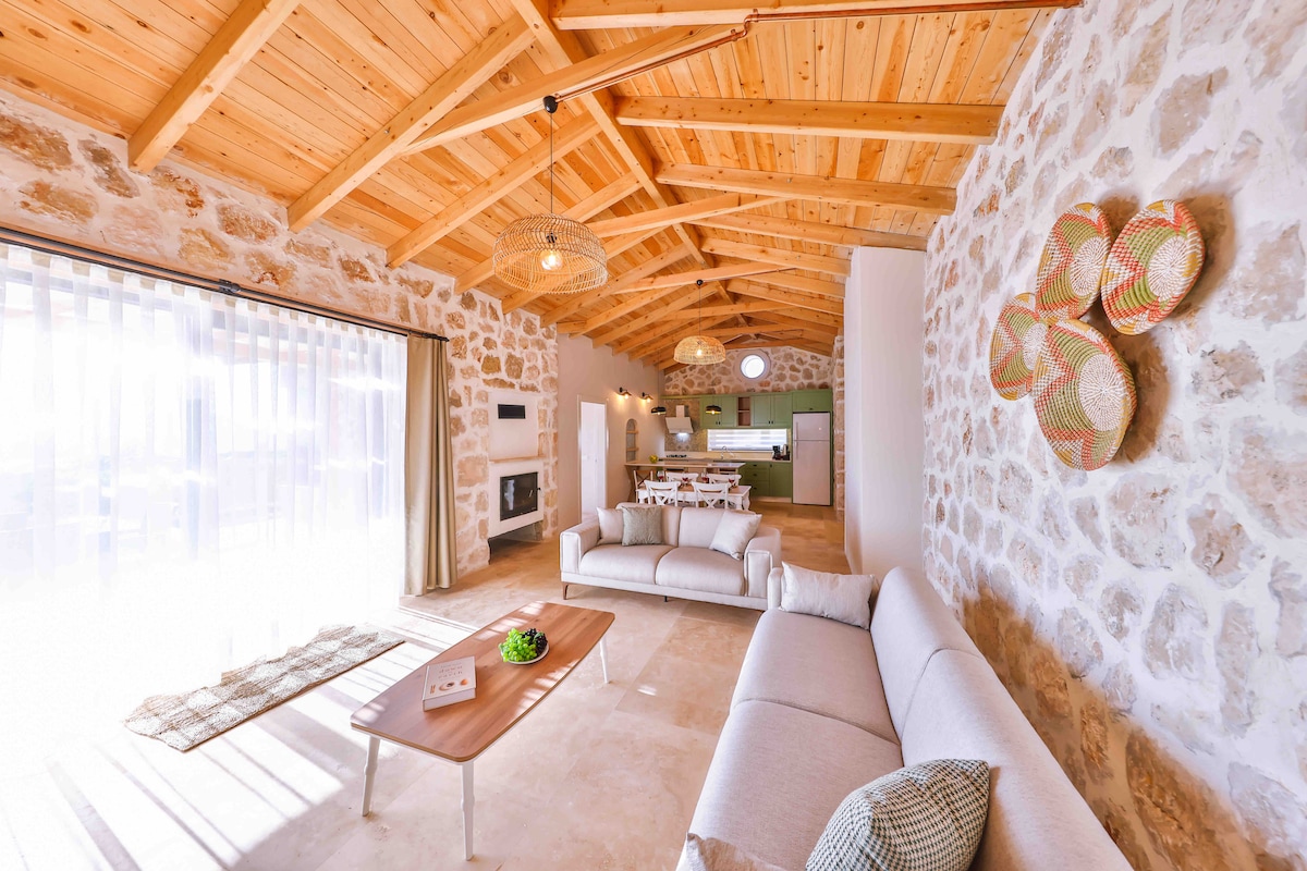 A spacious living area features a combination of stone walls and wooden beams, creating a rustic yet modern ambiance. Soft, light-colored sofas are arranged around a central coffee table. Large glass doors allow natural light to illuminate the space, leading to an outdoor view.