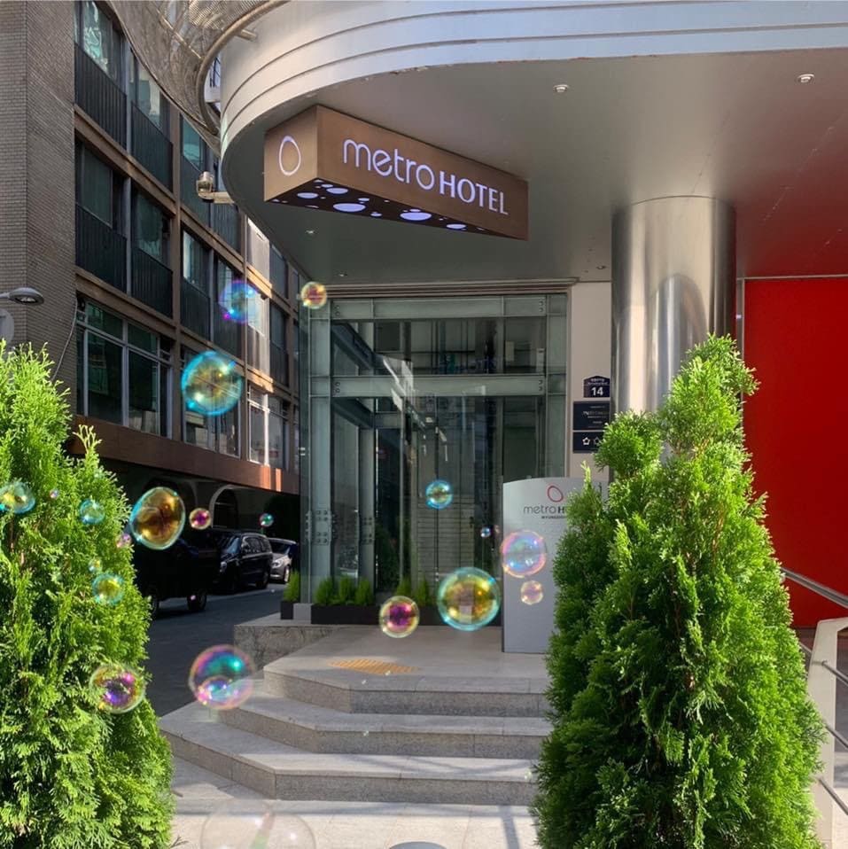 The entrance of Metro Hotel is framed by neatly trimmed greenery, leading up to a set of steps. A modern building facade is displayed alongside the hotel signage, indicating its name clearly. The atmosphere appears inviting and accessible.