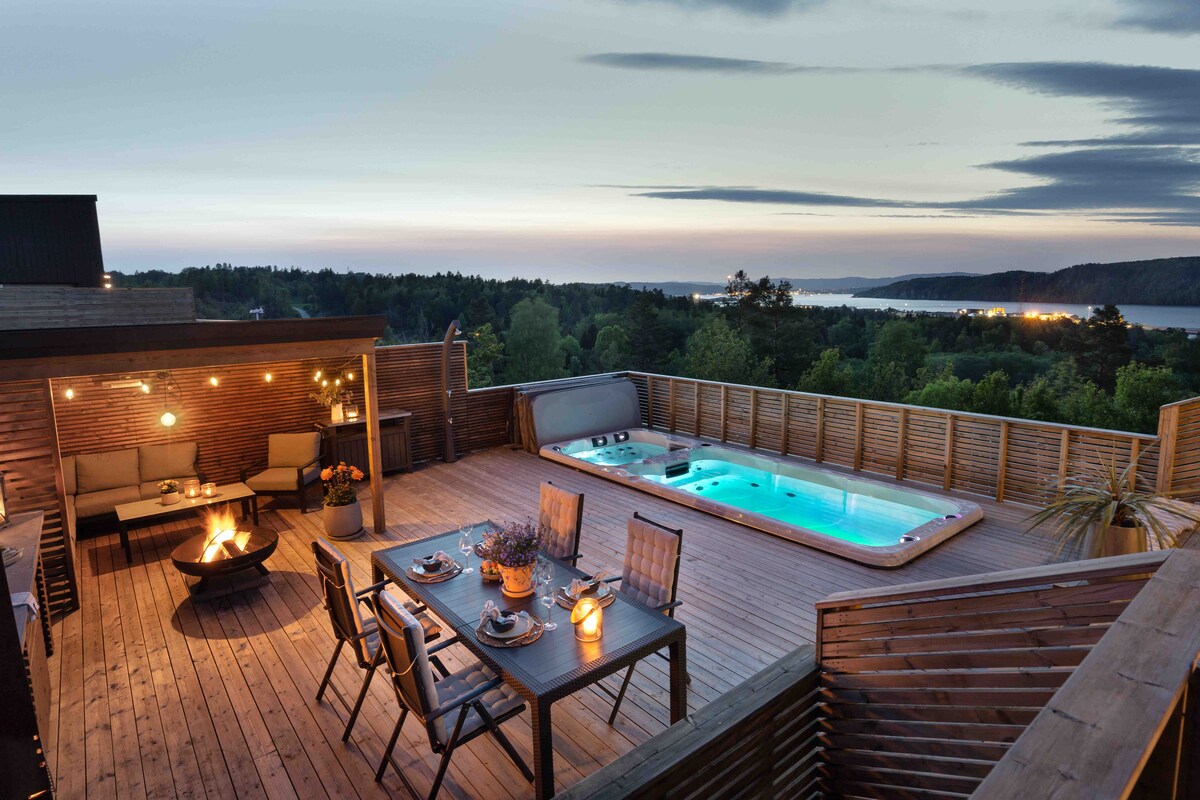 An outdoor deck area features a dining table set with chairs, surrounded by soft lighting. A hot tub is positioned nearby, with a scenic view of the landscape and water in the background, illuminated by the evening sky.