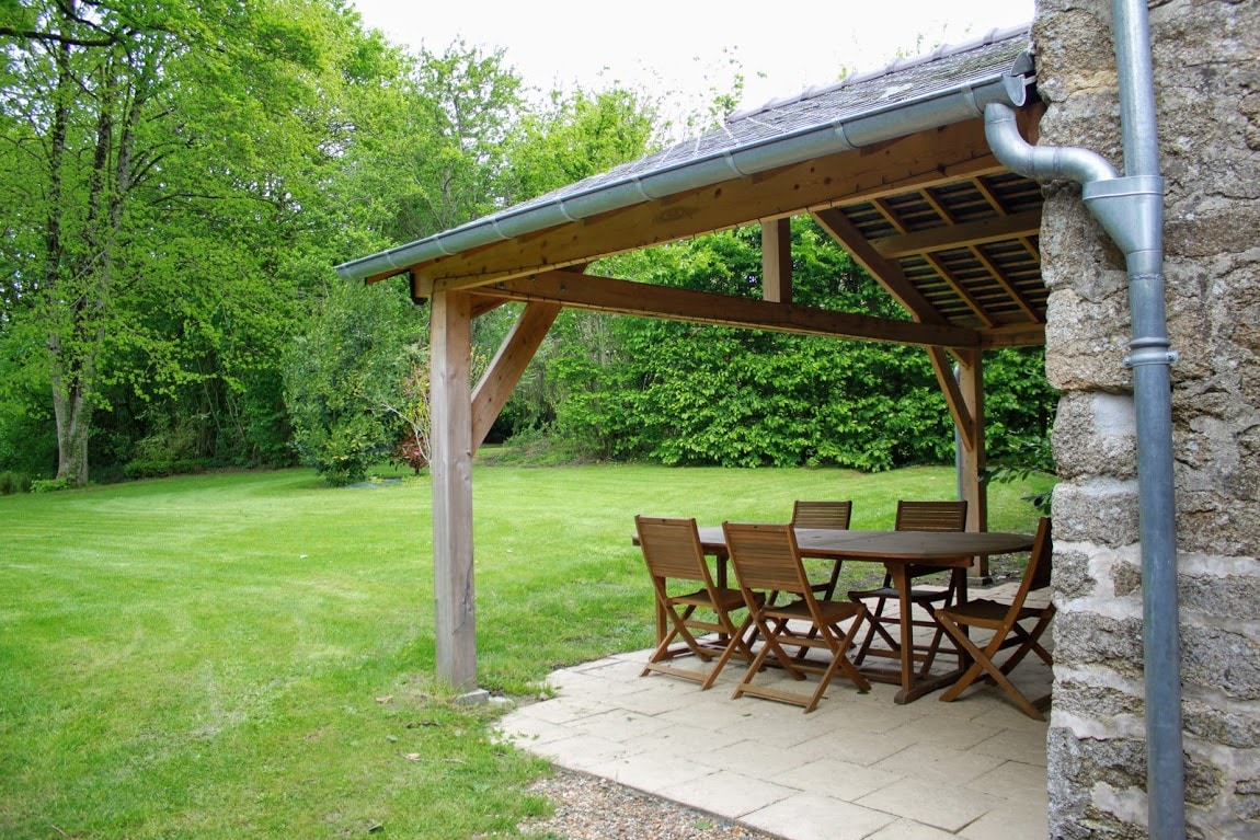An outdoor terrace features a wooden pergola supported by sturdy posts. A rectangular table is surrounded by six folding chairs, set on a stone-paved area. Lush green grass is visible in the expansive private garden, bordered by trees and bushes for added privacy.
