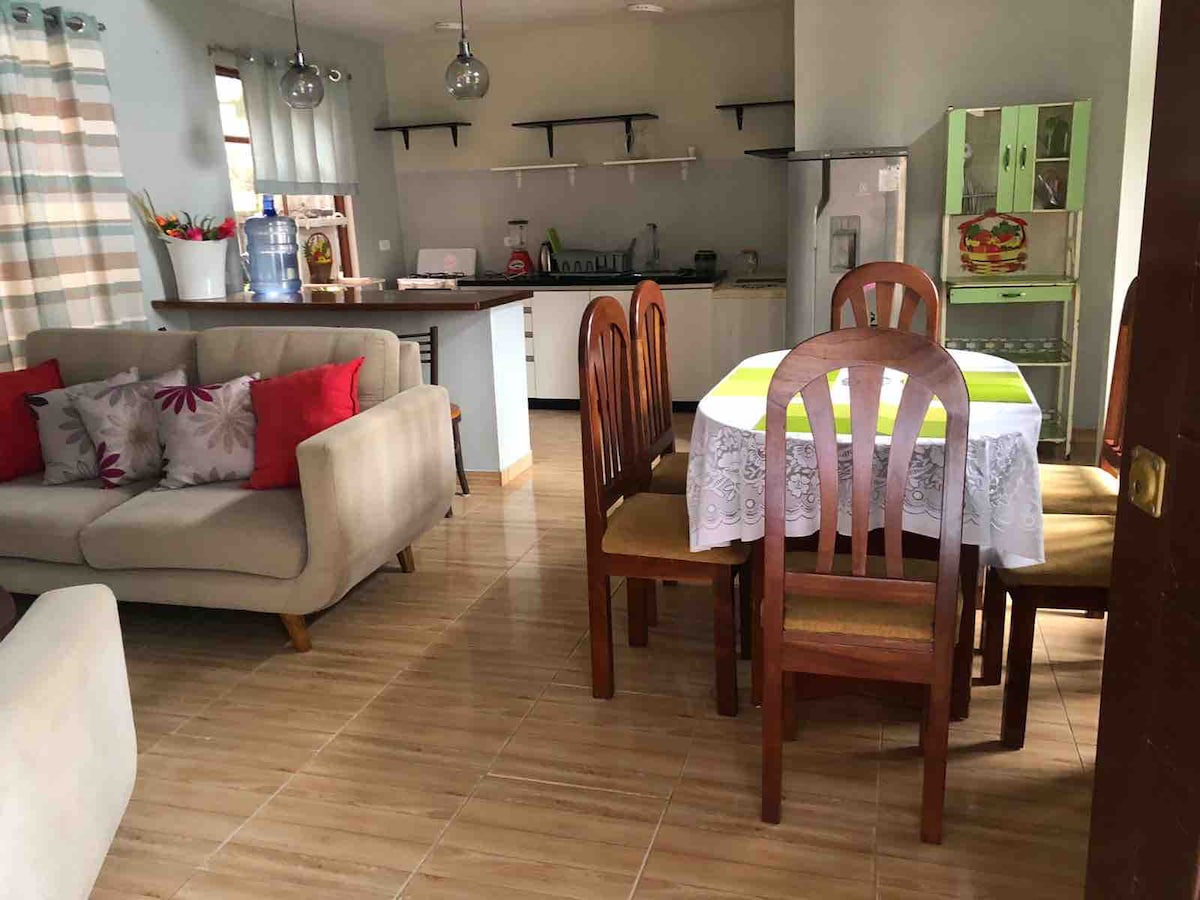 An open-plan living and dining area features a light-colored sofa paired with patterned cushions. A wooden dining table with six chairs sits next to the kitchen, which includes modern appliances and ample counter space. Natural light brightens the space through large windows.