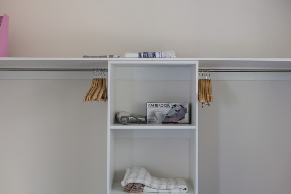 A well-organized closet space is featured, with wooden hangers neatly arranged and a stack of towels on a shelf. A small appliance is placed on the bottom shelf, accompanied by additional linens. The clean, modern design emphasizes functionality.