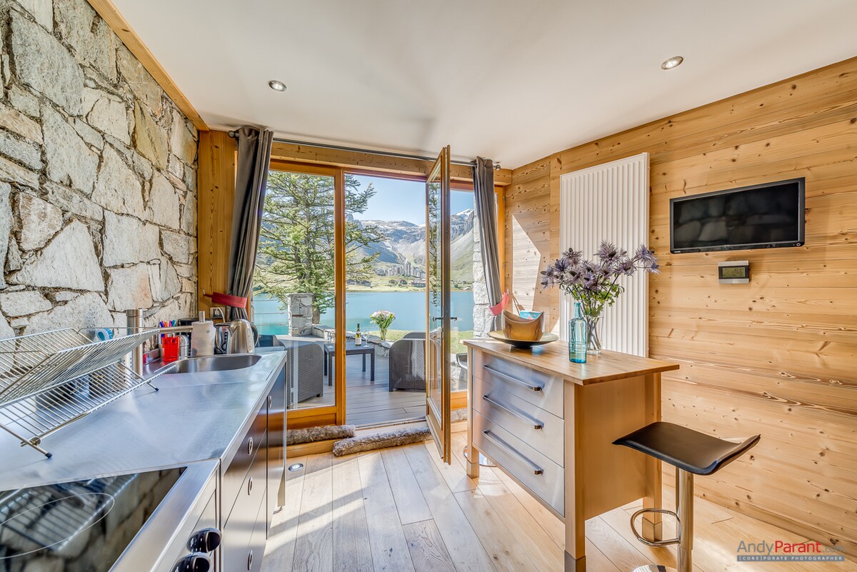 A modern kitchen area features sleek appliances along a wooden countertop. Large glass doors open to a terrace, allowing natural light and views of the lake and mountains. A small dining table with a flower centerpiece complements the wooden accents of the space.