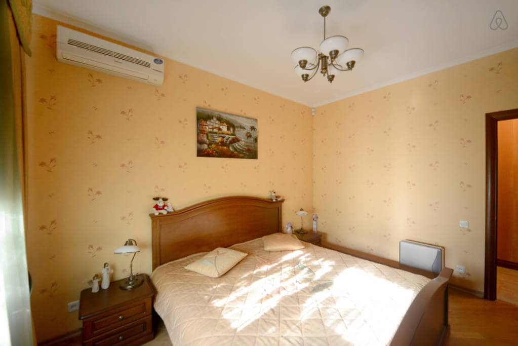 The bedroom features a king-size bed with a light beige quilt and decorative pillows. Two bedside tables with lamps are positioned beside the bed. A wall-mounted air conditioning unit is visible. A floral artwork decorates the wall above the bed, complemented by soft yellow wallpaper.