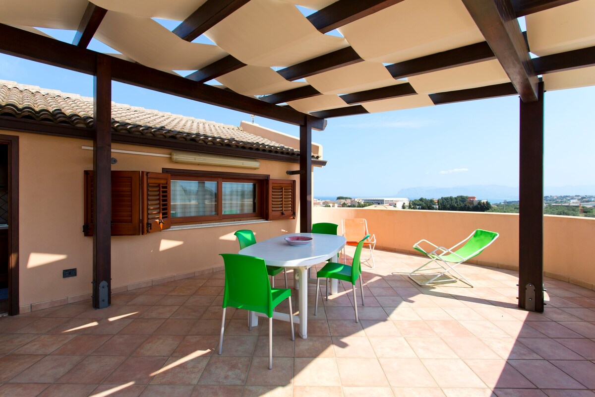 A spacious terrace features a dining table surrounded by six green chairs. A reclining lounge chair is positioned nearby, and shaded by white awnings. The view includes distant mountains and a clear blue sky.