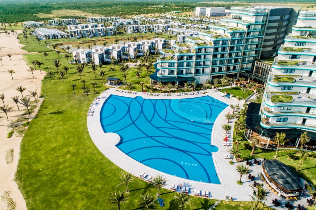 An aerial view showcases the expansive outdoor swimming pool, shaped with flowing patterns, surrounded by lush greenery and palm trees. The resort buildings, designed with a modern aesthetic, are visible in the background alongside well-maintained grounds, creating a spacious and inviting atmosphere.