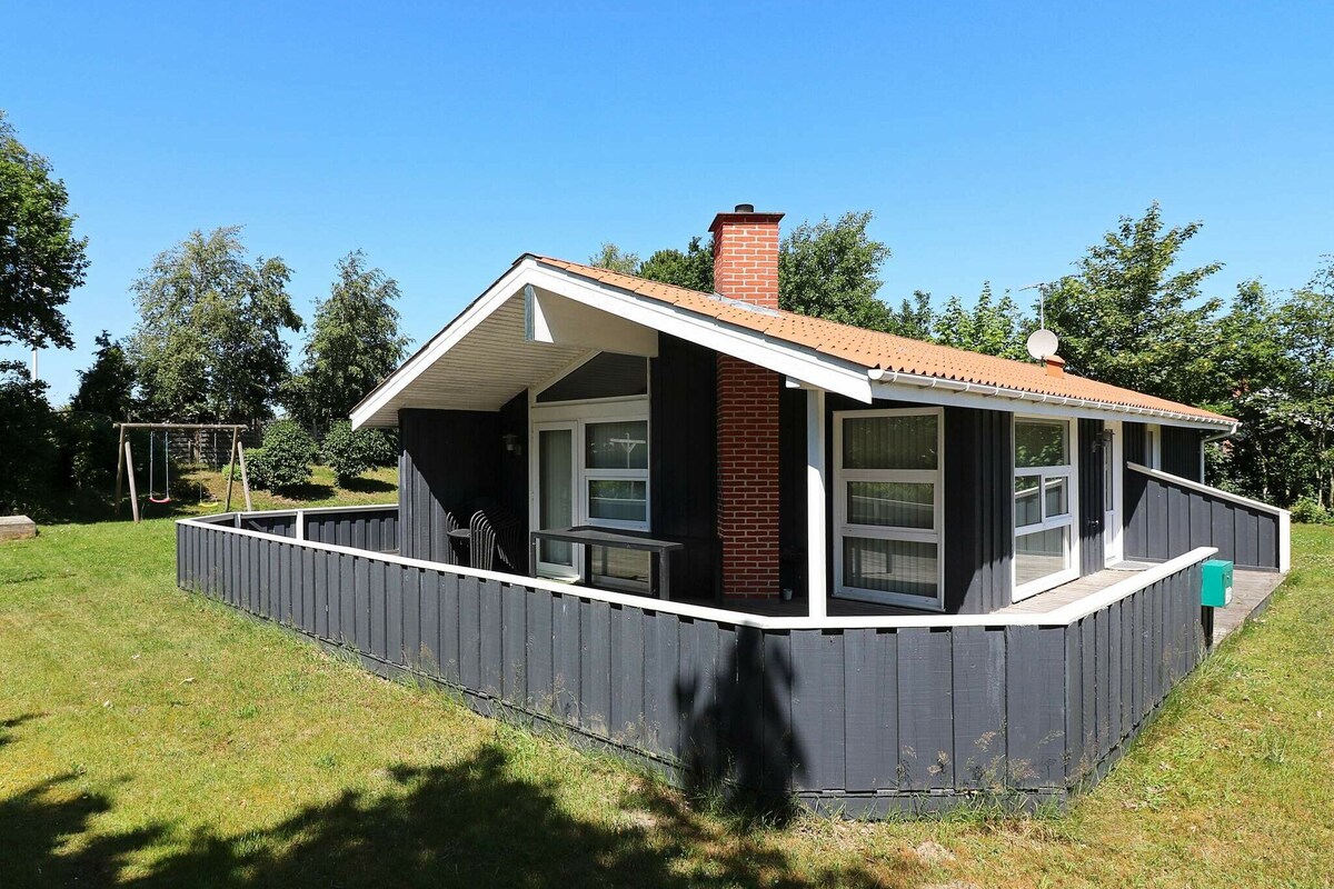 A holiday home is set amidst a grassy landscape, featuring a sloped roof and a prominent chimney. Large windows enhance natural light, while a shaded entryway and a swing set invite outdoor relaxation. Surrounded by trees, the house offers a peaceful setting.