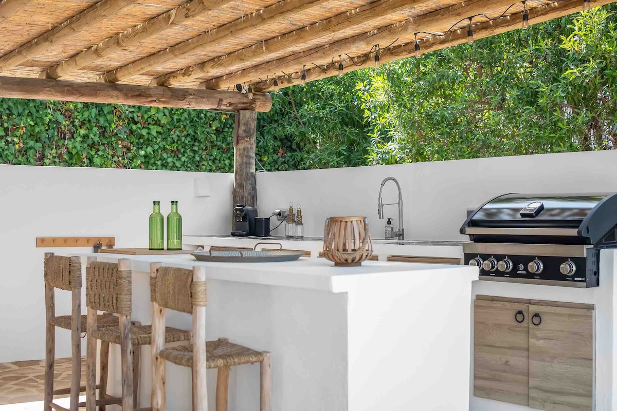 An outdoor kitchen area is highlighted by a spacious countertop and a modern grilling station. Natural materials are used throughout, with tall stools positioned at the counter. Lush greenery surrounds the space, adding to the inviting ambiance.