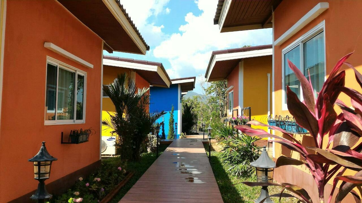 A pathway lined with colorful cottages leads through the resort, bordered by vibrant green plants and decorative lanterns. Blue skies and gentle clouds provide a serene backdrop, enhancing the inviting atmosphere of the space.