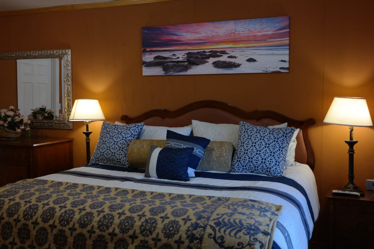 A spacious bed is adorned with patterned pillows and a coordinating throw blanket, set against a backdrop of warm red clay walls. Two bedside lamps provide soft lighting, while a landscape painting captures a serene coastal scene above the headboard.