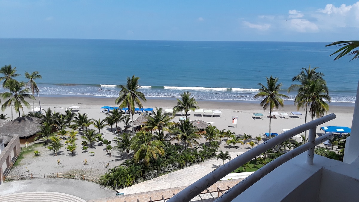 Vast ocean views are showcased from the balcony, featuring gentle waves lapping at the sandy shoreline. Palm trees line the beach, complemented by beach umbrellas and lounge chairs. A clear blue sky adds to the serene coastal ambiance.