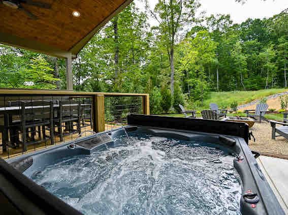 A hot tub filled with bubbling water is set on a wooden deck, surrounded by greenery. In the background, a dining table is visible under a covered area, while several outdoor chairs are positioned nearby on a gravel surface.