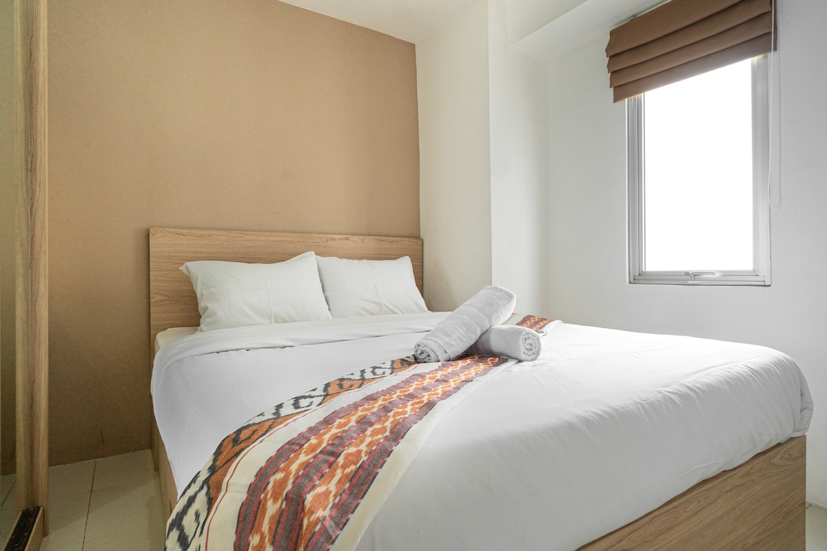 A cozy bedroom features a spacious queen-size bed with crisp white linens and decorative accents. A large window provides natural light, while a set of brown blinds offers privacy. A plush towel is neatly rolled and placed on the bed.