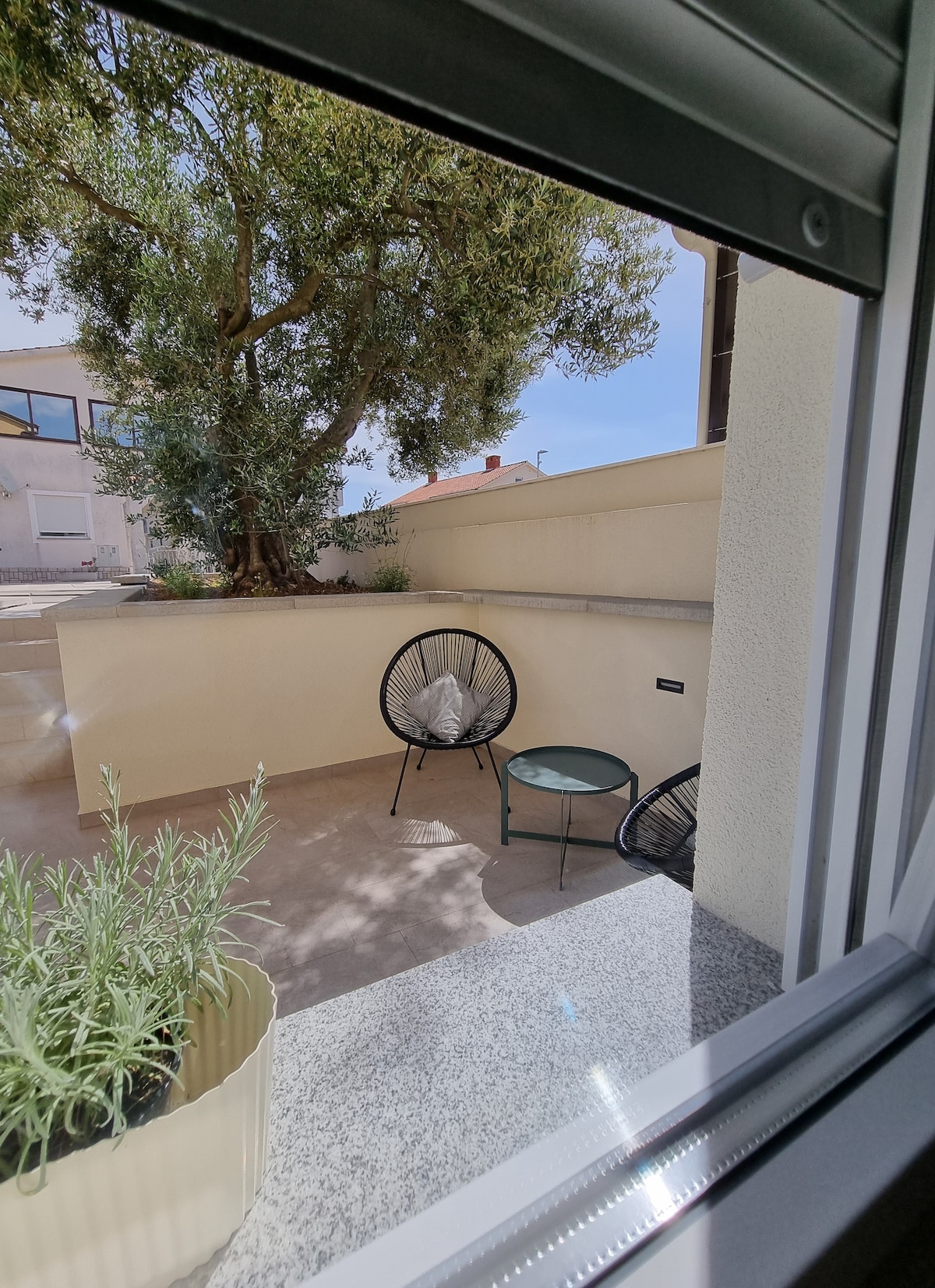 An outdoor seating area is visible through a window, featuring two black woven chairs positioned on a stone terrace. A small green accent table is beside one chair, and a planter with lavender adds a touch of greenery to the space. Sunlight brightens the scene.