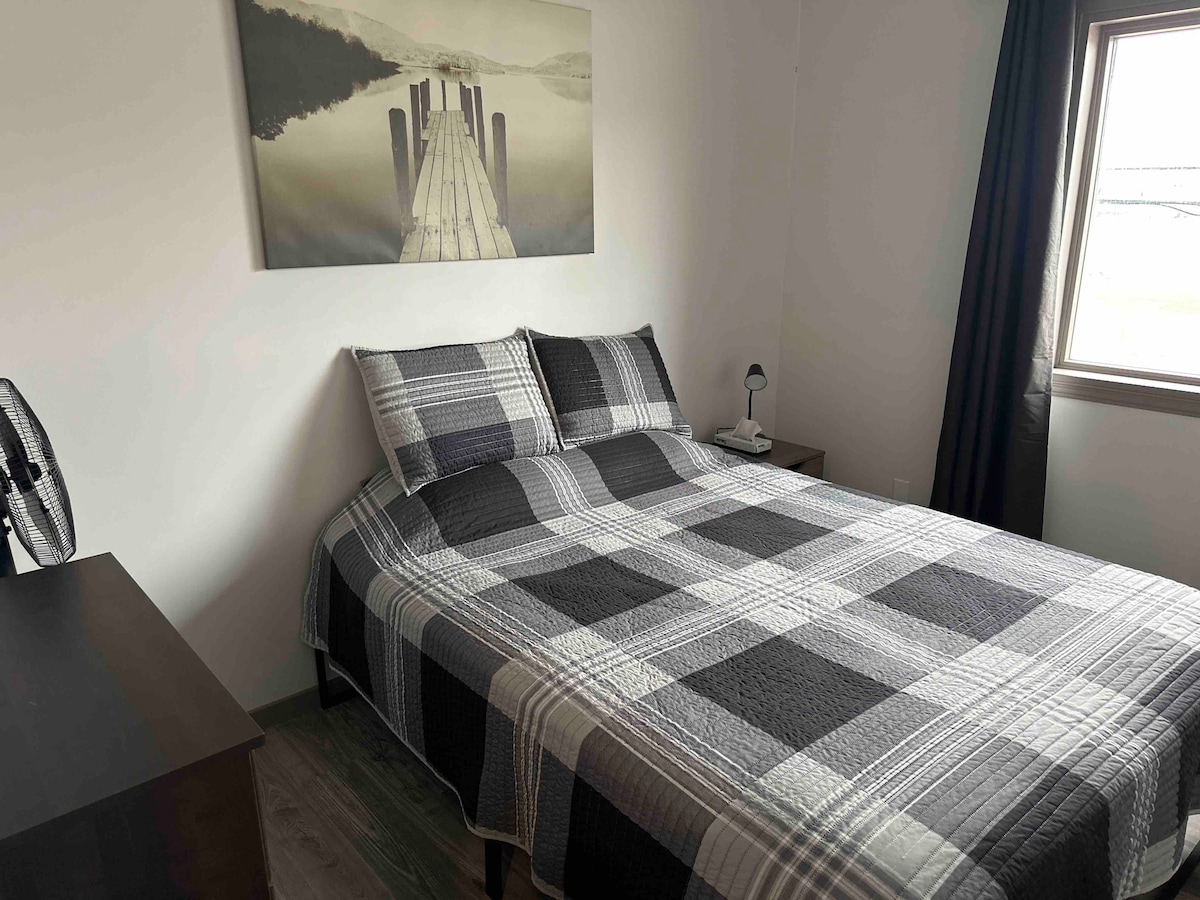A cozy sleeping area features a spacious bed dressed in a checkered blanket. A bedside table with a lamp is situated next to the bed. Natural light filters through a window adorned with dark curtains, while a fan sits nearby to enhance comfort.