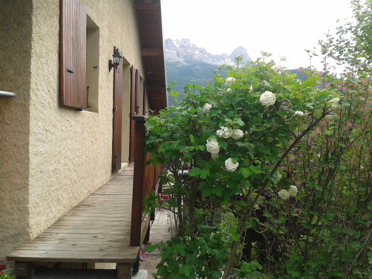 A wooden pathway leading to the entrance of a beige house is flanked by lush green foliage and blooming white flowers. Majestic mountains rise in the background, providing a natural and serene backdrop to the scene.